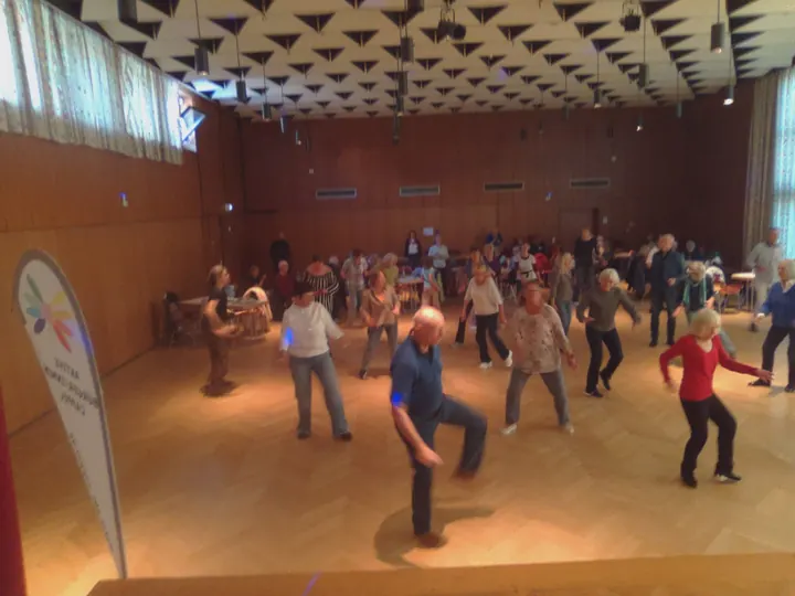 Teilnehmende üben gemeinsam Tanzbewegungen; im Vordergrund wird ein Knie angehoben.